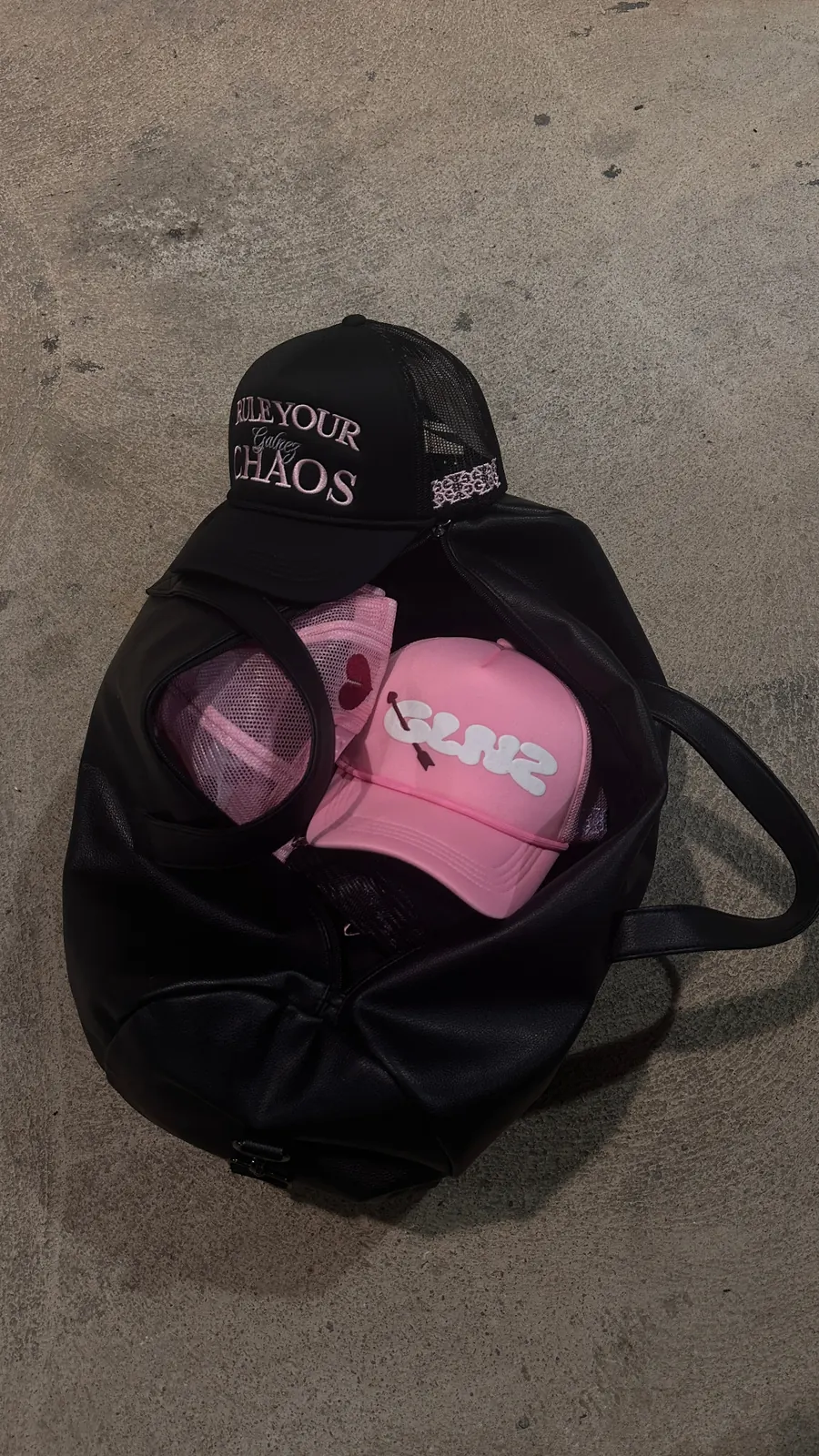 Black GALNEZ 'Rule Your Chaos' trucker cap on a duffle bag with pink caps inside